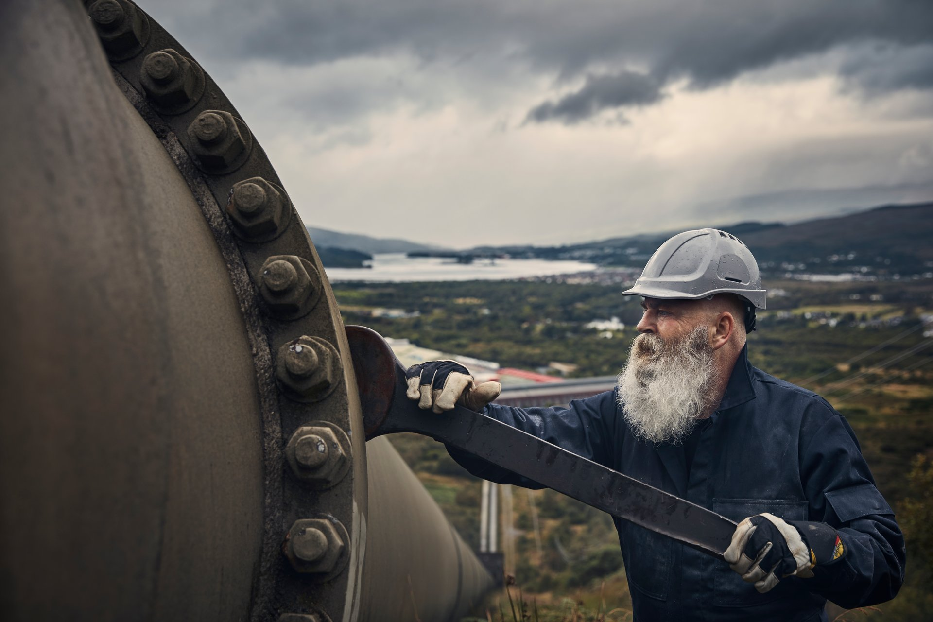 Man with a beard wearing a hard hat and gloves, standing next to a large pipe with a scenic background.