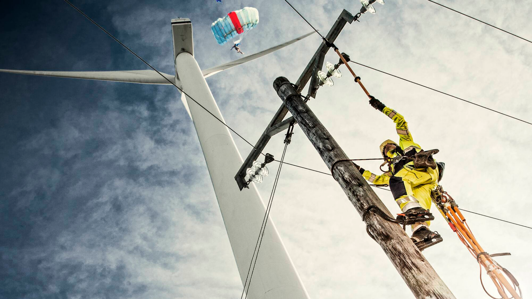 Person in yellow safety jacket working on a power line with a blue sky and white clouds in the background.