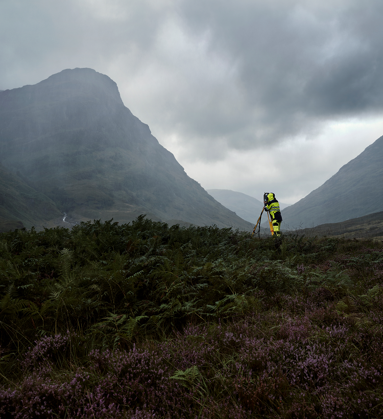 Person in high-visibility jacket standing in a mountainous landscape with misty mountains and overcast sky.