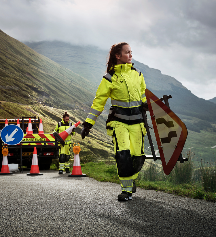 Person in high-visibility safety gear walking on a road with traffic cones and a sign, mountains in the background.