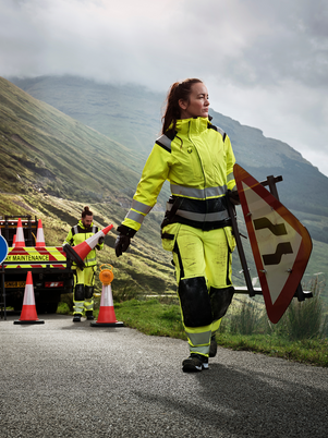Person in high-visibility safety gear walking on a road with traffic cones and a sign, mountains in the background.